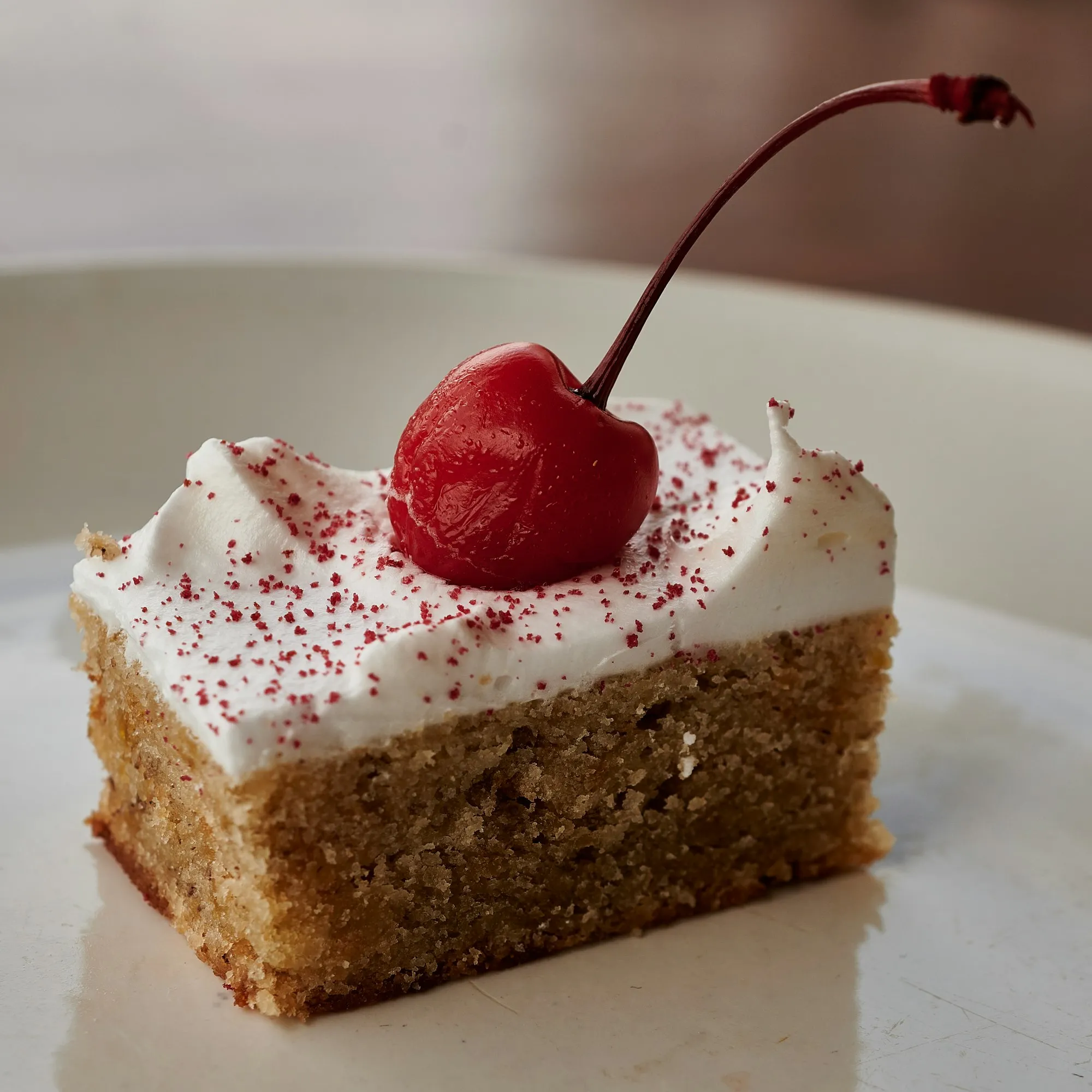 A square of sponge cake with whipped cream and a glossy cherry on top