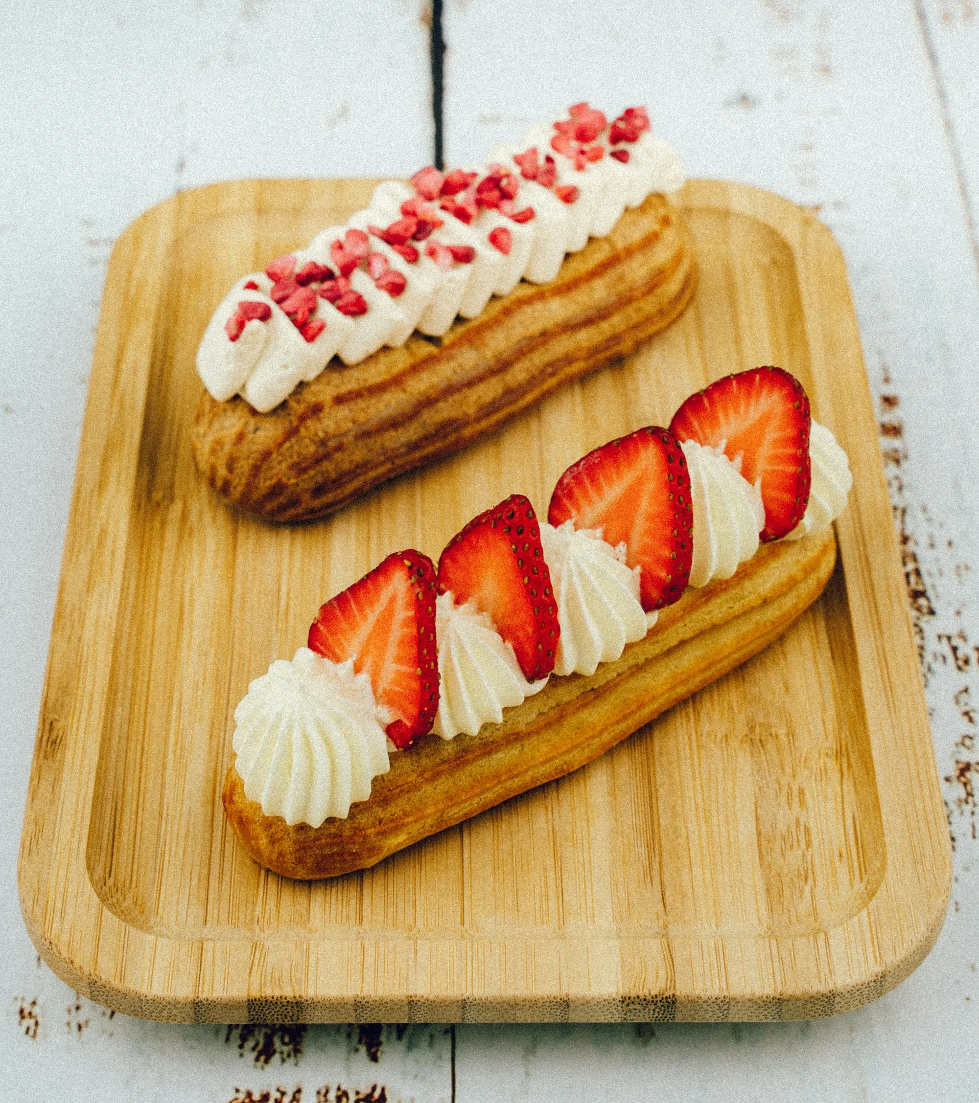 Two strawberry éclairs topped with cream and freeze-dried fruit on a wooden board